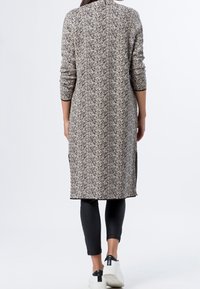 Cardigan a lunghezza ginocchio, a motivo in crema e nero, con maniche lunghe, fessure laterali e design a collo alto. Indossato su leggins neri e scarpe.