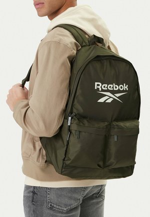 Mężczyzna w beżowej kurtce i białej bluzie z kapturem niesie oliwkowo-zielony plecak Reebok z dwoma przednimi kieszeniami na zamek.