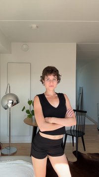 T-shirt noire court et shorts. Cheveux courts et bouclés. Chambre avec un sol en bois, une table ronde, une plante et une lampe moderne. Couleur des murs neutre.
