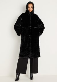 Abrigo de felpa negro con capucha, con grandes botones, un exterior suave y un ribete en contraste. Diseño de ajuste oversized con una longitud media.