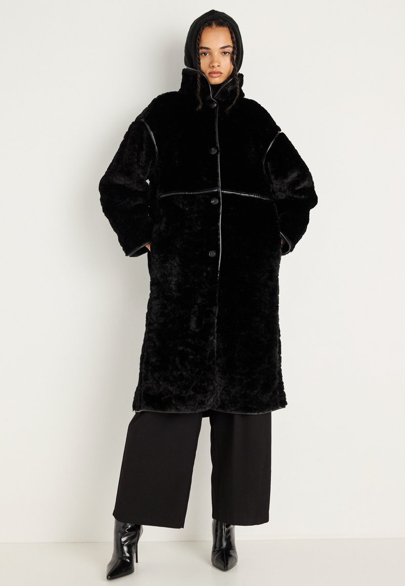 Abrigo de felpa negro con capucha, con grandes botones, un exterior suave y un ribete en contraste. Diseño de ajuste oversized con una longitud media.