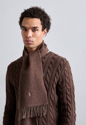 Man met krullend haar die een bruin kabelgebreide trui en een bijpassende bruine wollen sjaal met franjes draagt, tegen een effen grijze achtergrond.