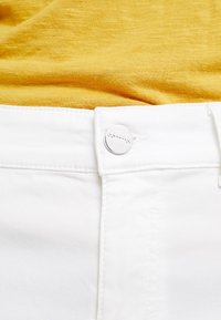 Jean en denim blanc à texture lisse, avec un bouton en métal argenté à la taille. Un tissu jaune est partiellement visible au-dessus du jean.