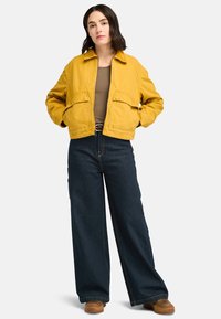 Yellow cropped jacket with two front pockets, worn over a brown top. Paired with wide-leg dark denim pants and brown shoes.
