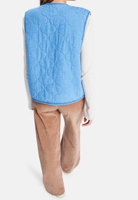 Gequilteste lichtblauwe bodywarmer van denimachtig materiaal met afgeronde patronen. Gecombineerd met beige corduroy broek met brede pijpen.