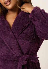 Accappatoio viola in morbido tessuto peloso, con un finish strutturato, dotato di colletto sciallato e cintura in vita per una vestibilità sicura. Decorato con maniche ampie.