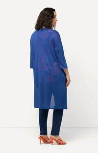 Modrý průhledný dlouhý kardigan s třičtvrtinovými rukávy, zkombinovaný s tmavými džíny a oranžovými sandály na podpatku. Texturovaná, lehká látka.