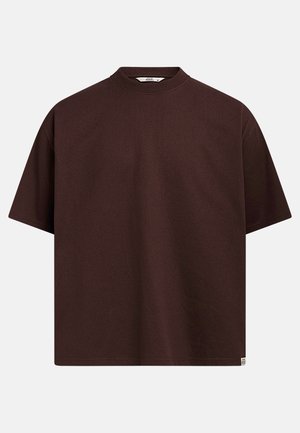 T-shirt à manches courtes, marron foncé, oversized, avec un col rond et une petite étiquette en tissu blanche au bas de l'ourlet.