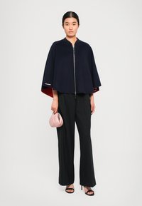 Cape en mélange de laine bleu marine avec un intérieur rouge, fermetures à glissière à l'avant et accents latéraux. Associée à un pantalon plissé noir et une pochette rose.