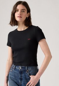 T-shirt noir à manches courtes avec un col rond et une texture côtelée. Présente un logo Levi's rouge sur la poitrine. Associé à un jean en denim bleu.