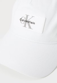 Witte pet van katoen, met een rechthoekig logopatch waarop "Calvin Klein" en "CK" in zilveren letters staan. Glad oppervlak, gebogen klep.