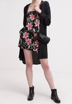 Femme portant une combishort fleurie noire sans bretelles, un long gilet noir ouvert, des bottines noires et portant un petit sac noir avec une bandoulière chaîne.