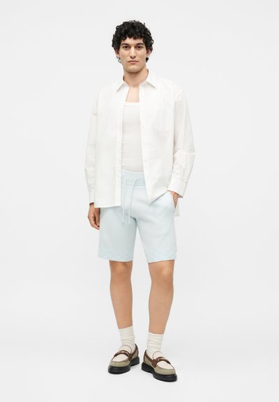 Άνδρας φοράει λευκό ανοιχτό πουκάμισο πάνω από λευκό φανελάκι, ανοιχτά μπλε σορτς, κρεμ κάλτσες και διχρωματικά μοκασίνια, στέκεται με το ένα χέρι στην τσέπη.