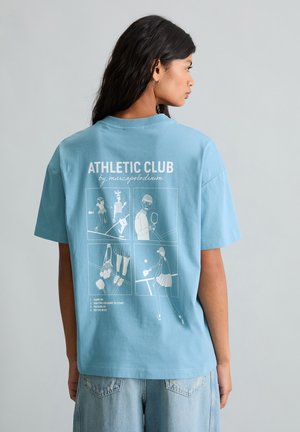 Frau in blauem T-Shirt mit der Aufschrift "ATHLETIC CLUB" und Cartoon-Tennisszenen auf der Rückseite, trägt hellblaue Jeans und steht vor einem schlichten Hintergrund.