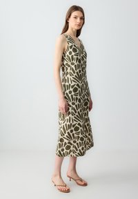 Mouwloze jurk in beige met groene abstracte bladrand, V-hals, vloeiende silhouette, valt onder de knie, gecombineerd met beige haksandalen.