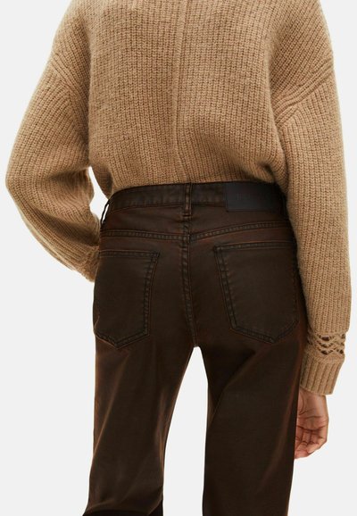 Pull en tricot marron avec un motif texturé, associé à un jean marron foncé avec poches arrières et une étiquette à la taille.