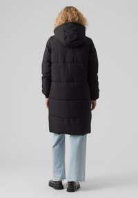 Svart dunjakke med hette, med en quiltet design og lange ermer. Kombinert med lyseblå vide jeans og svarte sko.