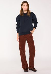 Donna che indossa un hoodie blu navy, pantaloni in velluto a coste marroni e mocassini marroni con nappine, in piedi con una mano in tasca su uno sfondo uniforme.