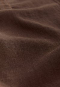 Brązowa lniana tkanina o miękkiej fakturze, z subtelnymi wzorami splotu i delikatnym układem, ukazująca swoją lekkość i przewiewność.