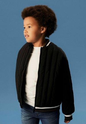 Dijete kovrčave kose koje nosi bijelu majicu, plave traperice i crnu prošivenu jaknu stoji ispred plave pozadine, gledajući ulijevo.