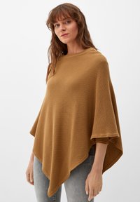 Poncho marrone, strutturato, con un design morbido e drappeggiato, maniche ampie e un orlo che forma un triangolo. Abbinato a jeans grigi aderenti.