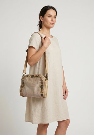 Femme en robe beige à manches courtes tenant un sac à main beige avec des accents métalliques ronds décoratifs.