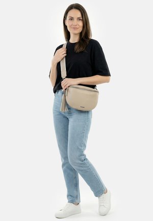Woman wearing black t-shirt, light blue jeans, white sneakers, holding beige crossbody bag with tassel against plain background.