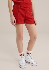 Červené textúrované šortky s elastickým pásom, spárované s bielymi vysokými teniskami a červenobielymi ponožkami po členky.