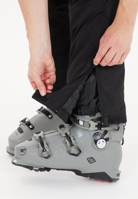 Harmaat hiihtokengät säädettävillä soljilla, tekstuuripinta sivuilla ja musta housunlahje, jossa on vetoketju ja Velcro-suljenta, osittain avattu.