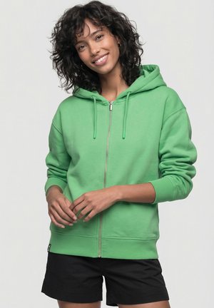 Frau mit lockigem Haar, die lächelt, trägt einen grünen Kapuzenpullover mit Reißverschluss und schwarze Shorts, steht vor einem schlichten hellen Hintergrund.