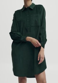 Zielona sukienka koszulowa z sztruksu, z zapięciem na guziki, długimi rękawami i kieszonką z przodu. Lekko oversize'owy krój z zaokrąglonym dołem.