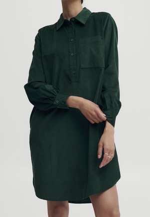 Abito camicia in corduroy verde con colletto a bottoni, maniche lunghe e una tasca frontale. Vestibilità leggermente oversize con orlo curvo.