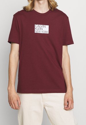 Osoba w bordowej koszulce Calvin Klein New York i kremowych spodniach, stojąca na tle jednolitego, jasnego tła.