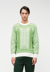 Pull en tricot vert avec un motif texturé représentant un terrain de tennis en blanc. Manches et ourlet en rib d'un blanc éclatant. Associé à un pantalon vert foncé.