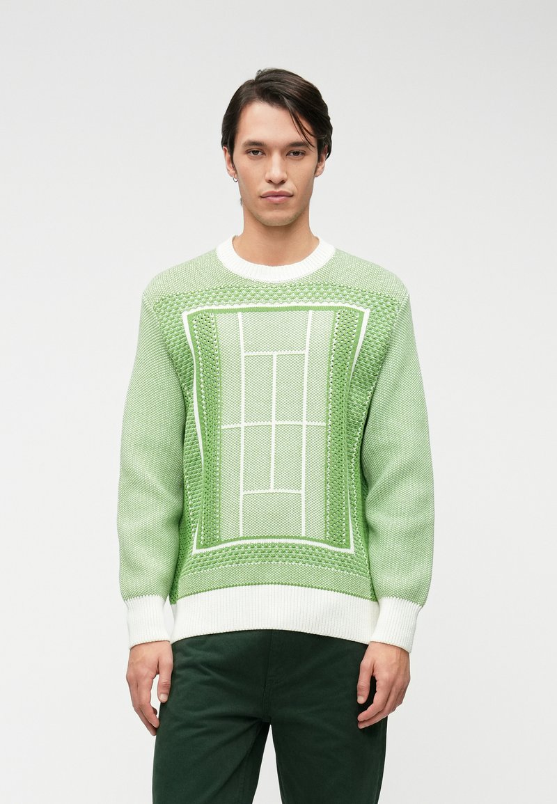 Pull en tricot vert avec un motif texturé représentant un terrain de tennis en blanc. Manches et ourlet en rib d'un blanc éclatant. Associé à un pantalon vert foncé.