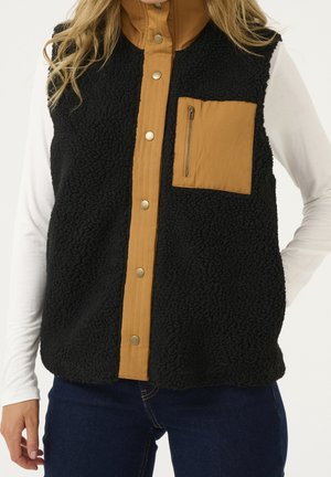 Kobieta ubrana w czarną kamizelkę z polaru sherpa z brązową patką na guziki i zapinaną na zamek kieszenią na piersi, na białą bluzę z długim rękawem i ciemne jeansy.