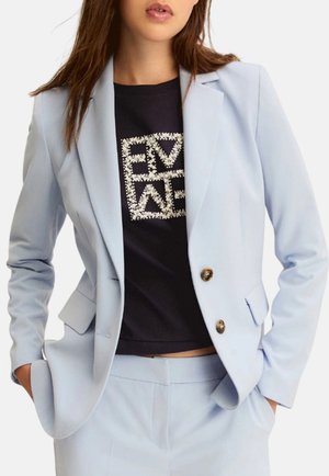 Donna con completo azzurro chiaro con due bottoni e top nero decorato con lettere di strass, mani in tasca.