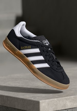 Baskets Adidas Gazelle noires avec des bandes blanches et une semelle en gomme flottant au-dessus d'une surface en pierre sur fond sombre.