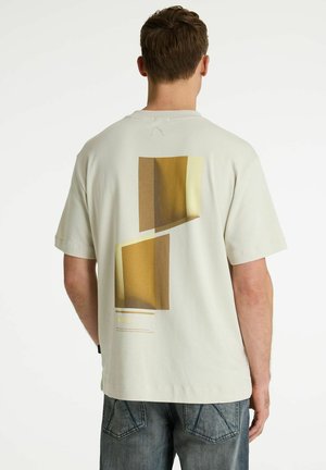 Man draagt een gebroken witte korte mouwen shirt met een geel-bruine abstracte geometrische print op de achterkant, gecombineerd met vervaagde blauwe spijkerbroek.
