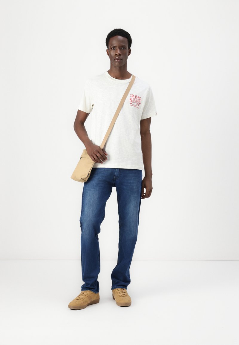 Helles graues T-Shirt mit rohem Grafik, blaue Jeans und beige Sneakers. Eine kleine beige Umhängetasche hängt von seiner Schulter.