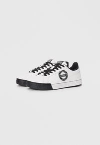 Balti odiniai sportbačiai su juodais akcentais, turintys apvalią logotipo etikėtę, juodas raištelius ir tekstūruotą juodą gumos pado paviršių.