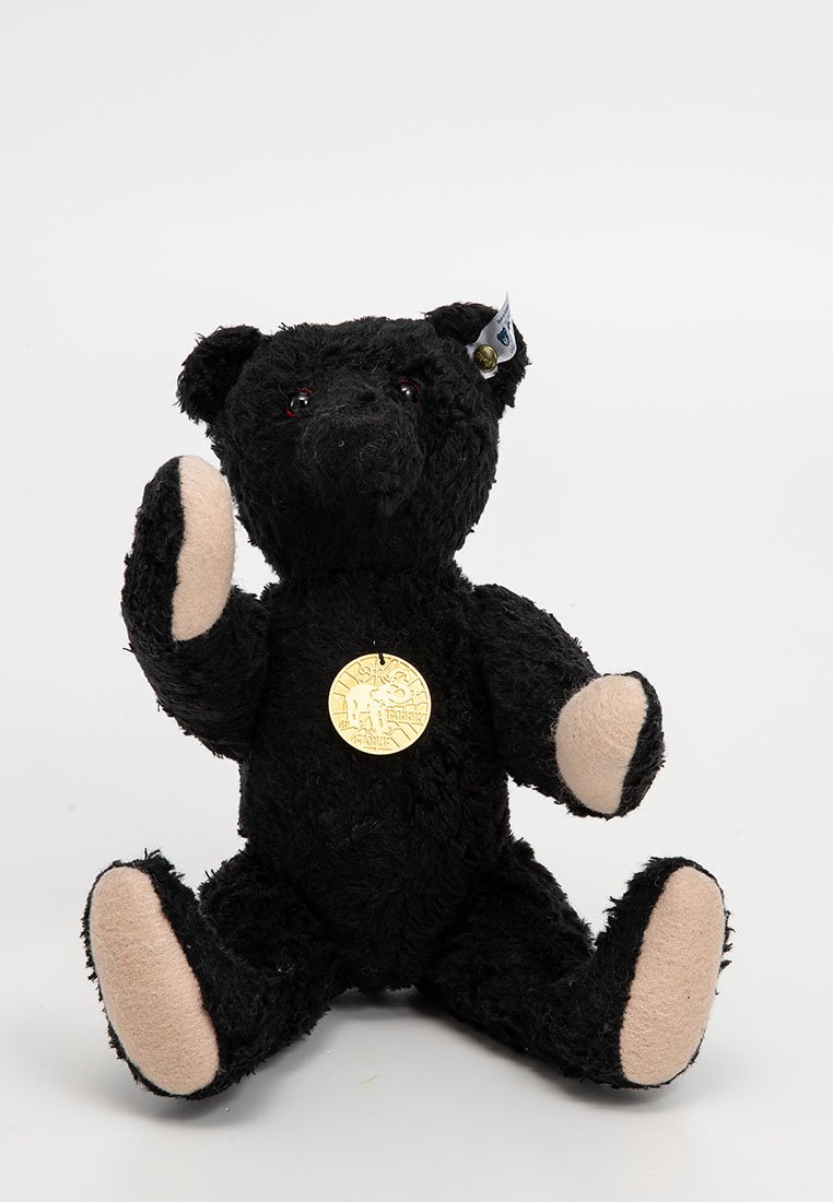 Schwarzer Teddybär mit beigen Pfotenunterseiten, einer goldenen Medaille auf der Brust und einem Anhänger im linken Ohr, der mit einem erhobenen Pfoten sitzt.