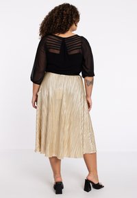 Zwarte doorschijnende blouse met streepmotief aan de achterkant, gecombineerd met een geplooide, metallic gouden rok en zwarte block-heeled sandalen.