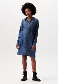 Vestido de camisa em denim azul escuro com mangas compridas, frente com botões, bolso de chapa, detalhe de cinta na cintura e bainha curva. Combinado com botins pretos.