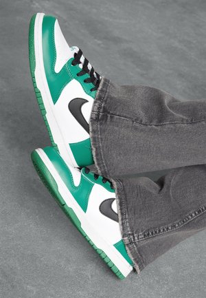 Baskets blanches et vertes avec logo noir, portées avec un jean gris sur fond gris, pieds croisés et légèrement soulevés du sol.