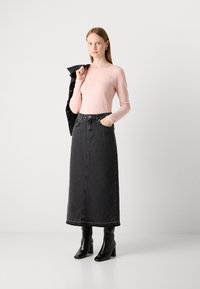 Žena s dugom plavom kosom nosi svijetloružičastu majicu dugih rukava, crnu traper suknju do poda s visokim strukom, crne gležnjače, dok drži crnu jaknu preko ramena.