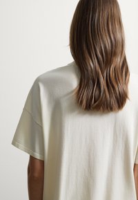 T-shirt color crema a maniche corte realizzato in tessuto morbido, con una vestibilità rilassata e dettagli delle cuciture visibili sul retro.
