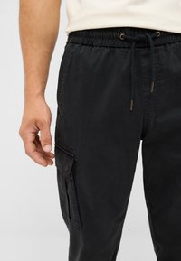 Man wearing black cargo pants with drawstring waist and flap pocket on left thigh, white shirt partially visible, hand relaxed by side.