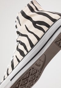 Vysoké plátěné tenisky mají béžový základ s černými pruhy zebry, bílé očka a texturovanou gumovou podrážku s vzorem.