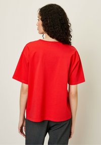 Raudona medvilninė marškinė su laisvu kirpimu, trumpomis rankovėmis ir apvaliu iškirpimu; vientisa spalva, lygi tekstūra, nematomi raštai ar aksesuarai.